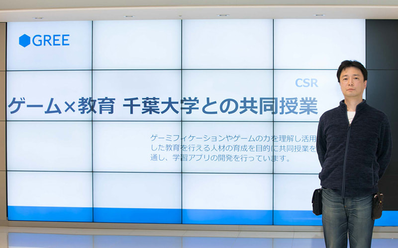 写真：大きなスクリーンに「ゲーム×教育 千葉大学との共同授業」の文字が映し出され、向かって右側に立つ小木曽健 氏